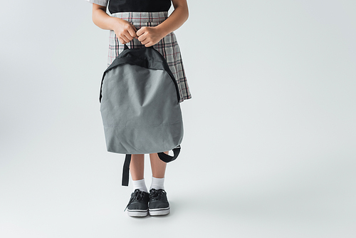 cropped view of schoolgirl in uniform holding backpack and standing on grey