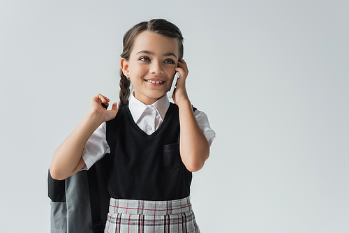 happy schoolgirl holding backpack and talking on smartphone isolated on grey