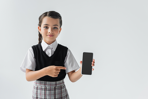 cute schoolgirl in uniform pointing with finger at smartphone with blank screen isolated on grey