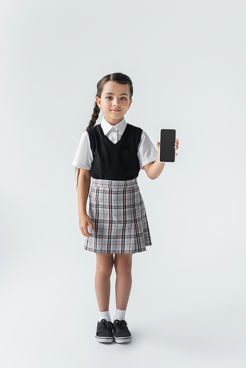 full length of cute girl in school uniform holding smartphone with blank screen on grey