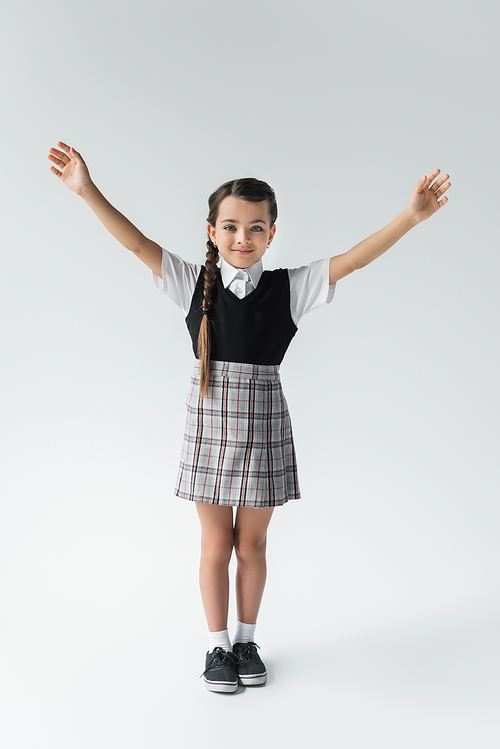 full length of excited schoolkid in uniform standing with outstretched hands on grey