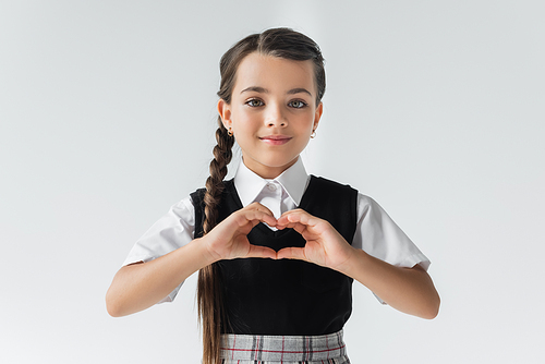 portrait of happy schoolgirl in white shirt and vest showing heart with hands isolated on grey