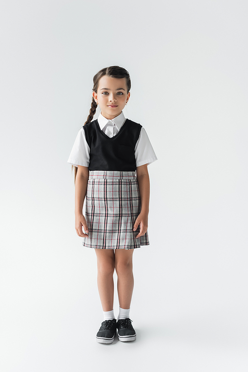 full length of cute schoolgirl in uniform standing and looking at camera on grey