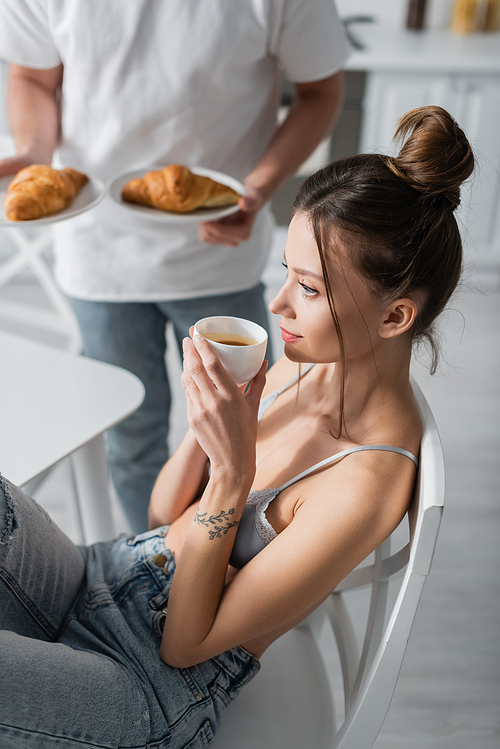tattooed woman holding cup near blurred man with croissants