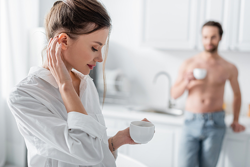 sensual young woman in white shirt holding cup of coffee near blurred and shirtless man on background