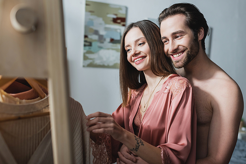 shirtless and happy man hugging tattooed girlfriend in pink silk robe choosing clothes