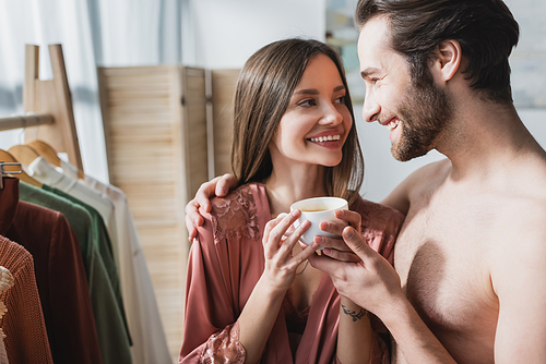 bearded and happy man hugging smiling girlfriend in pink silk robe holding cup of coffee