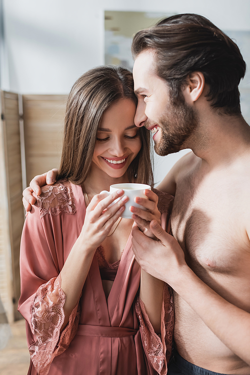happy man hugging smiling girlfriend in pink silk robe holding cup of coffee