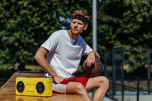 young basketball player sitting with ball near tape recorder and looking away