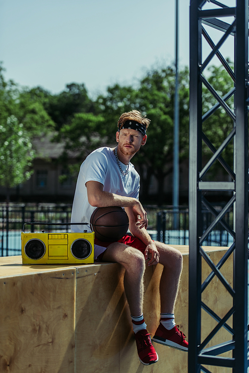 full length of man with ball sitting near boombox and looking at camera