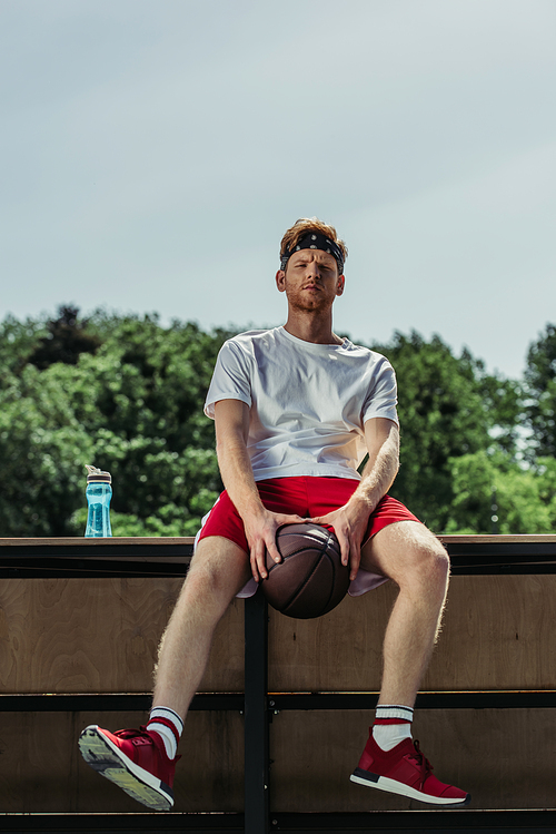 young sportsman holding ball while sitting near sports bottle and looking at camera