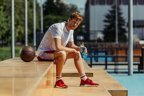 redhead man in sportswear sitting on stadium with ball and sports bottle