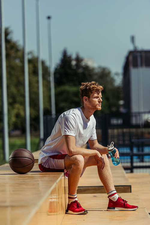 full length of man in red sneakers sitting with sports bottle near ball