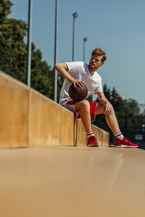 full length of man in red sneakers sitting on stadium with ball