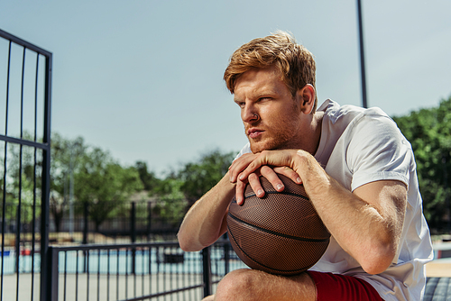 pensive basketball player sitting on stadium with ball and looking away