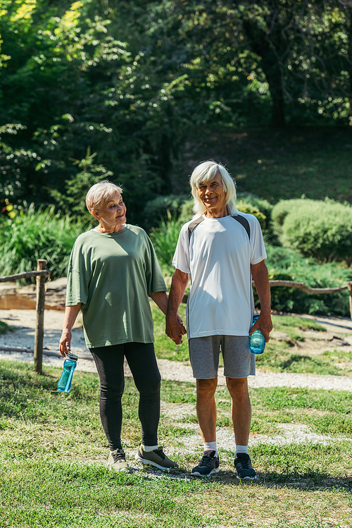 full length of happy retired couple in sportswear holding sport bottles and hands of each other in park