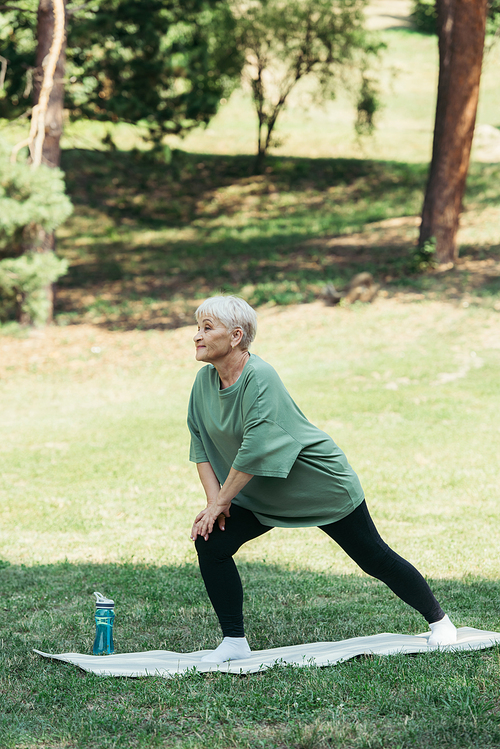 full length of senior woman with grey hair smiling and doing lunges on fitness mat in park