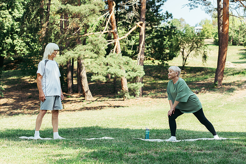 full length of senior man looking at wife doing lunges on fitness mat in park