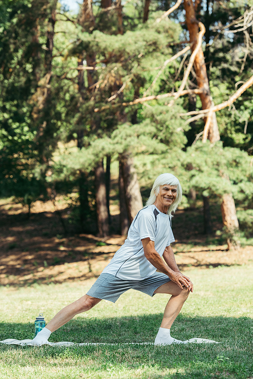 full length of senior man with grey hair smiling and doing lunges on fitness mat in park