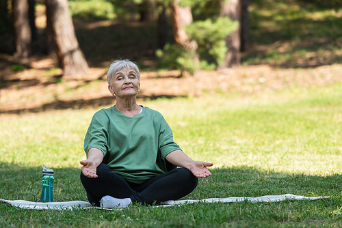 full length of senior woman with grey hair meditating on fitness mat in park