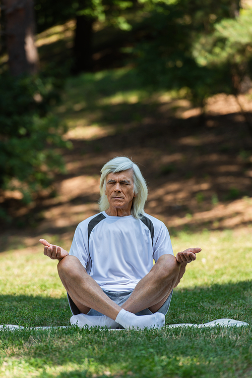 full length of senior man sitting in yoga pose and meditating on fitness mat in green park