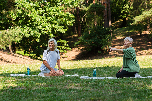 full length of senior woman pointing with finger near happy husband on fitness mat in green park