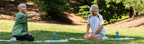 full length of senior woman pointing with finger near happy husband on fitness mat in green park, banner