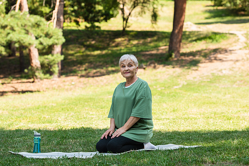 full length of senior woman with grey hair sitting on fitness mat in park