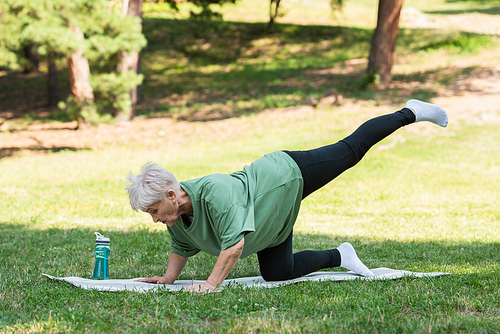 full length of senior woman with grey hair working out on fitness mat near sports bottle in park