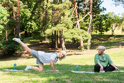 full length of senior woman in sportswear looking at husband exercising on fitness mat in green park