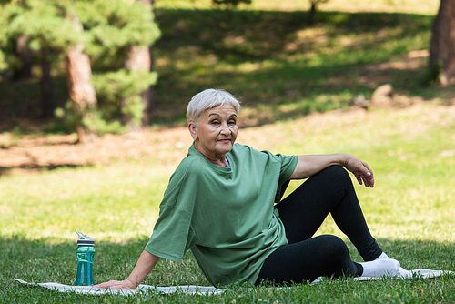 full length of senior woman sitting on fitness mat near sports bottle in park