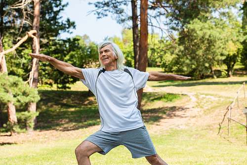cheerful senior man with grey hair smiling and working out with outstretched hands in park