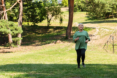 full length of senior woman with grey hair smiling and running on grass in park