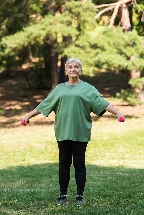 full length of cheerful senior woman with grey hair smiling and exercising with dumbbells in park