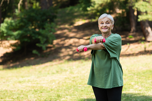 happy senior woman with grey hair smiling and exercising with dumbbells in park