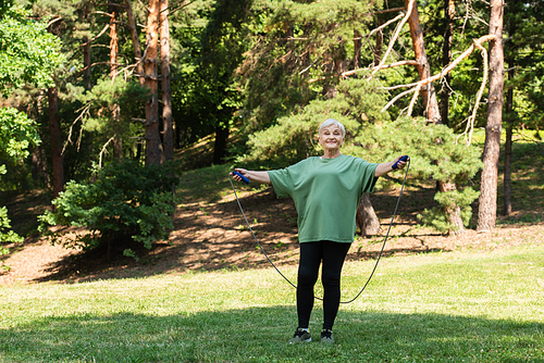 full length of senior woman with grey hair smiling and exercising with jumping rope in park