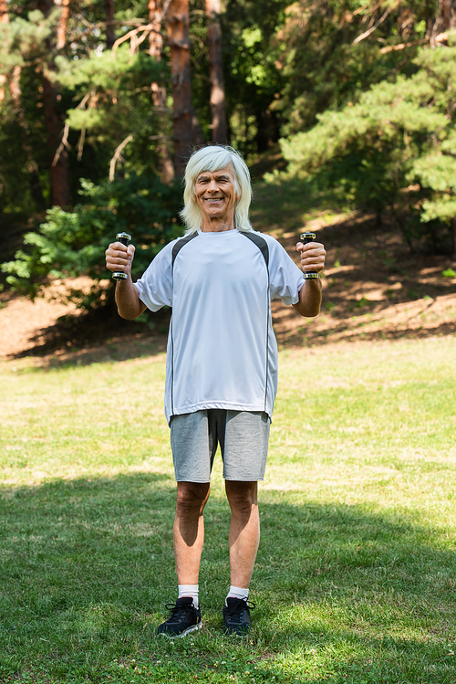 full length of happy senior man in sportswear working out with dumbbells in green park