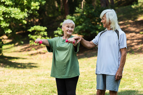 cheerful senior man in sportswear touching hand of wife working out with dumbbells in green park