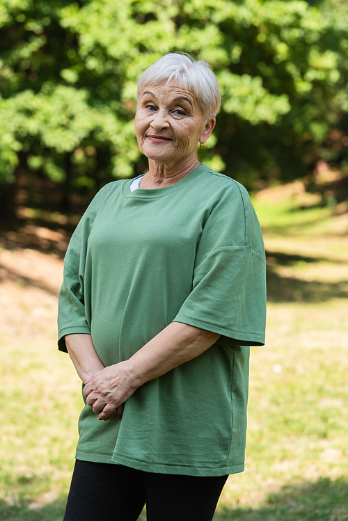 cheerful and retied woman with grey hair standing in green t-shirt in park