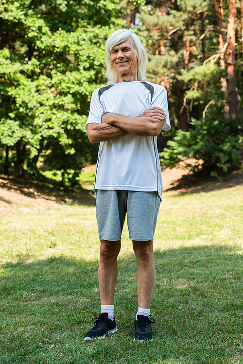 full length of happy senior man in sportswear standing with crossed arms in green park
