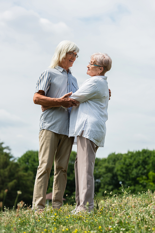 full length of happy senior husband and wife with grey hair hugging on green hill in summer