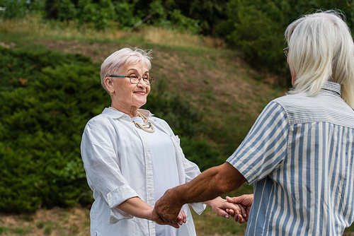 happy senior woman in glasses holding hands with husband in striped shirt in park