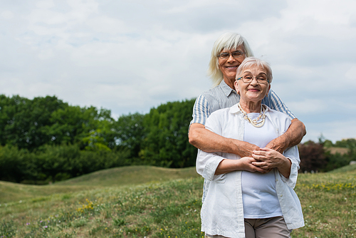 cheerful senior man in glasses hugging wife with grey hair and standing in park