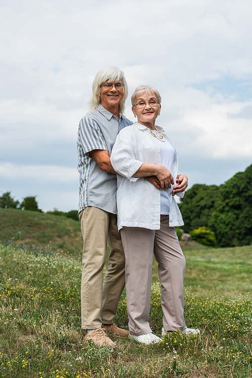 joyful senior man in glasses hugging happy wife with grey hair and standing on green hill