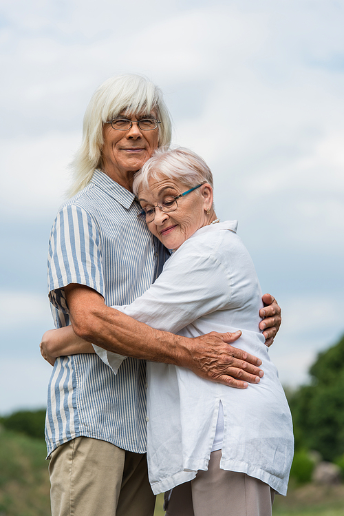 happy senior husband and wife with grey hair embracing against cloudy sky