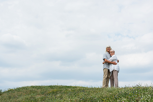 happy senior husband and wife with grey hair hugging and standing on green hill