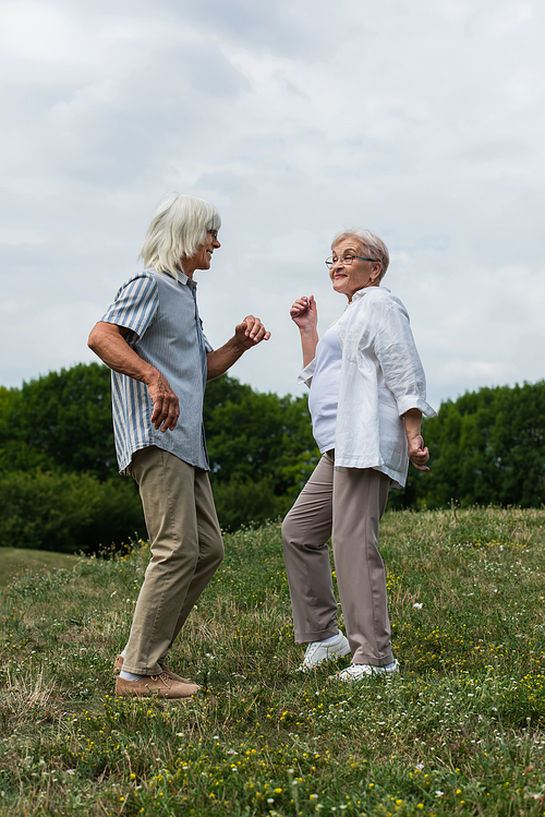 full length of happy retired couple in glasses dancing on green hill