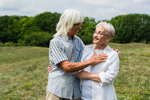 smiling senior couple in glasses hugging and looking at each other on green hill in summer