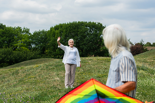 happy senior woman in casual clothes waving hand at husband with kite on green hill
