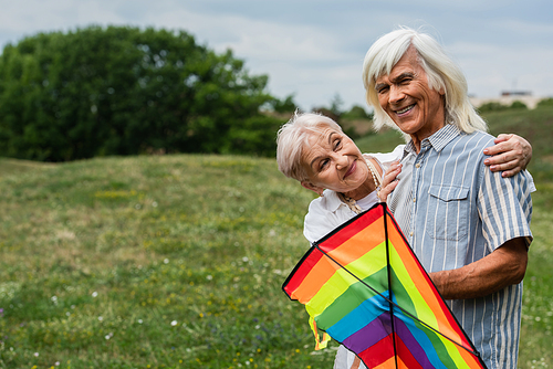 happy senior woman in casual clothes hugging husband with kite on green hill